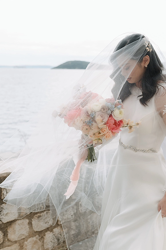 Bridal portrait of a bride holding bouquet with a long veil over her face, in a white dress by an ocean stone wall and sky