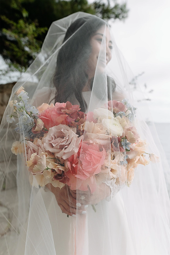 Bridal portrait of a bride holding bouquet, veil over her face, in a strapless wedding dress with trees and sky behind her