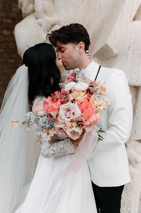 Wedding kiss portrait of bride and groom kissing, bride holding a bouquet in lace sleeves and veil, groom in white tux, stone wall backdrop