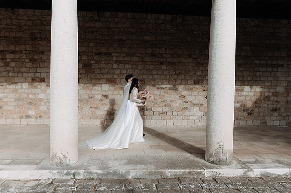 Couple portrait of bride and groom walking along a columned stone walkway, bride in lace sleeves with long veil holding bouquet