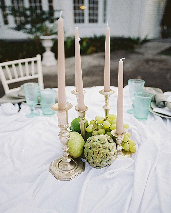 Wedding tablescape with taper candle centerpiece on a white tablecloth, brass candlesticks, grapes and pears, on an outdoor patio by a white facade