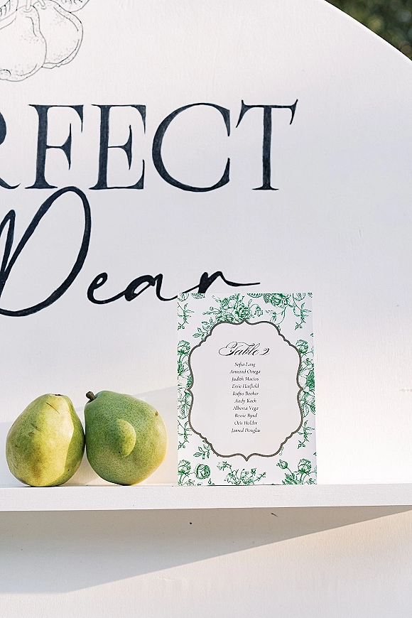 Wedding seating chart with wedding escort cards in calligraphy, paired with pears and floral illustration on a white backdrop with greenery