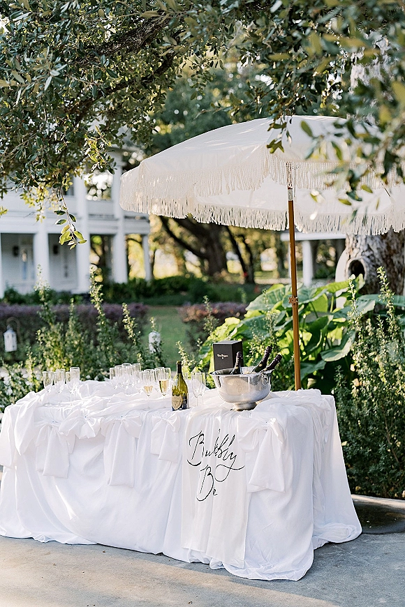 Champagne station setup with champagne flutes and bottles on a white linen table, accented by a fringe umbrella in a garden setting