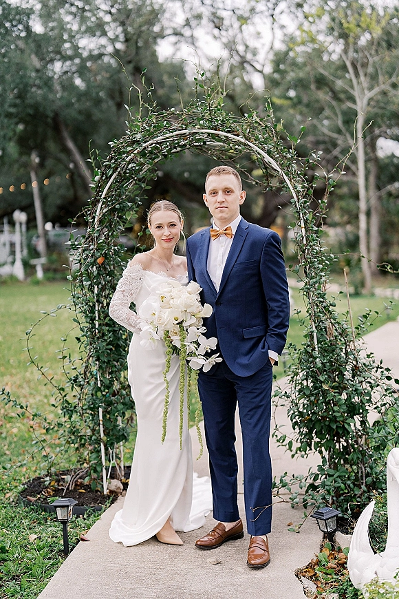 Couple portrait of bride and groom portrait under a vine-covered greenery arch, bride holding a cascading white orchid bouquet on a garden walkway