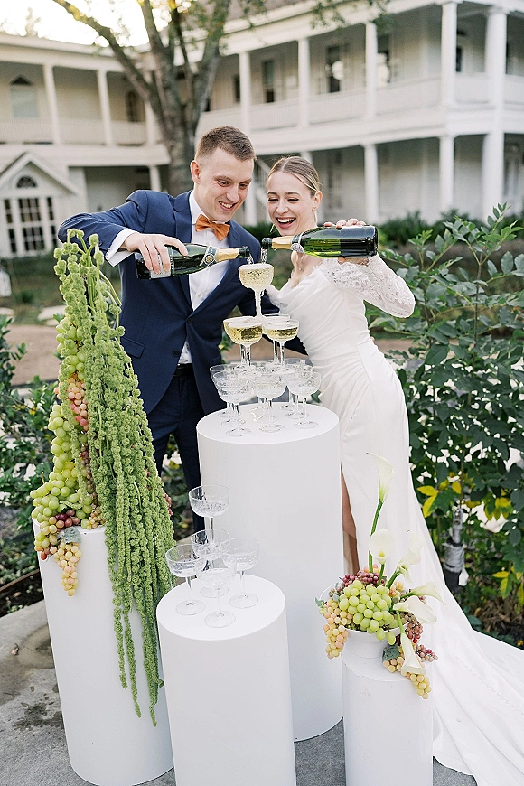 Champagne tower moment as bride and groom pour bubbly into coupe glasses on white pedestals, with calla lilies in a garden setting