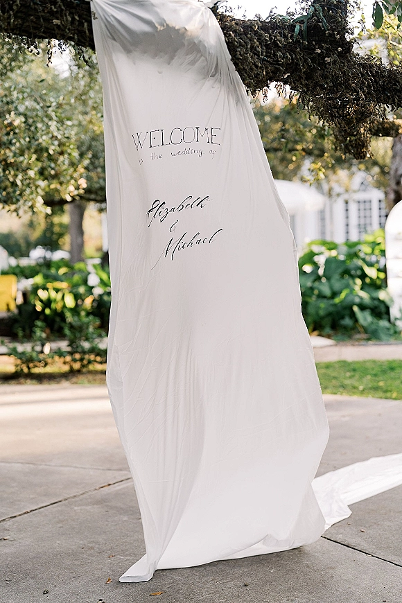 Wedding welcome sign with calligraphy lettering hanging from a mossy tree branch with fabric drape in a garden near a white building