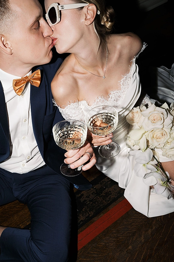 Wedding kiss portrait of bride and groom kissing while holding champagne coupes, bride in sunglasses and lace dress in dark interior
