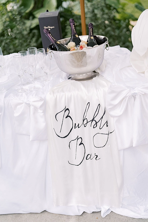 Wedding bubbly bar with champagne bar wedding sign, bottles in a silver ice bucket and flutes on a white draped table, roses against green foliage
