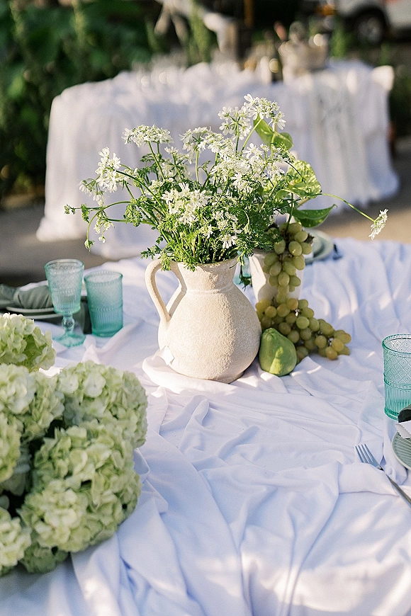 Wedding tablescape with white floral centerpiece in a ceramic pitcher, green grapes and pear, aqua glassware on a white tablecloth outdoors