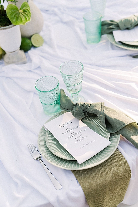 Wedding place setting with a sage green napkin and plate topped by a menu card, green glassware, silver flatware, and limes on white linen outdoors