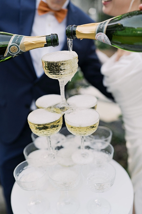 Champagne tower wedding pour as bride and groom tip bottles over stacked coupe glasses, bubbles spilling with greenery and guests blurred behind