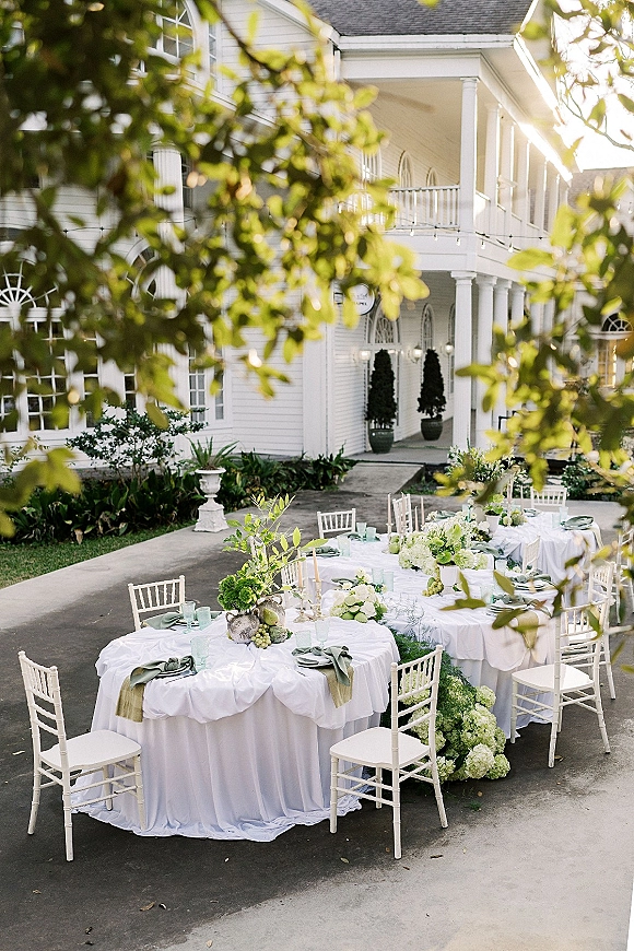 Outdoor reception tablescape with white linen round tables, hydrangea and greenery runners, taper candles, and fruit on an estate porch with string lights