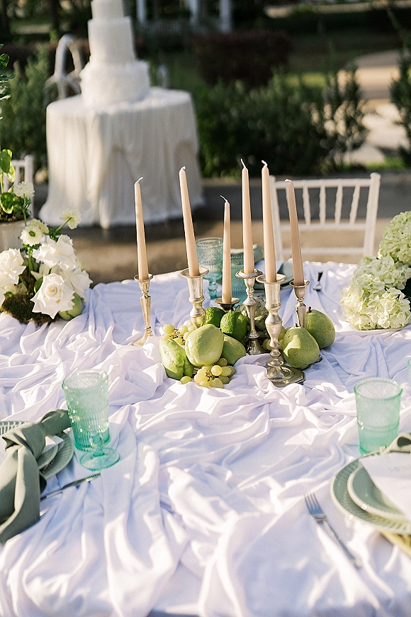 Reception tablescape with outdoor wedding table decor, white roses and hydrangeas, silver candlesticks, green pears, and taper candles on a garden patio