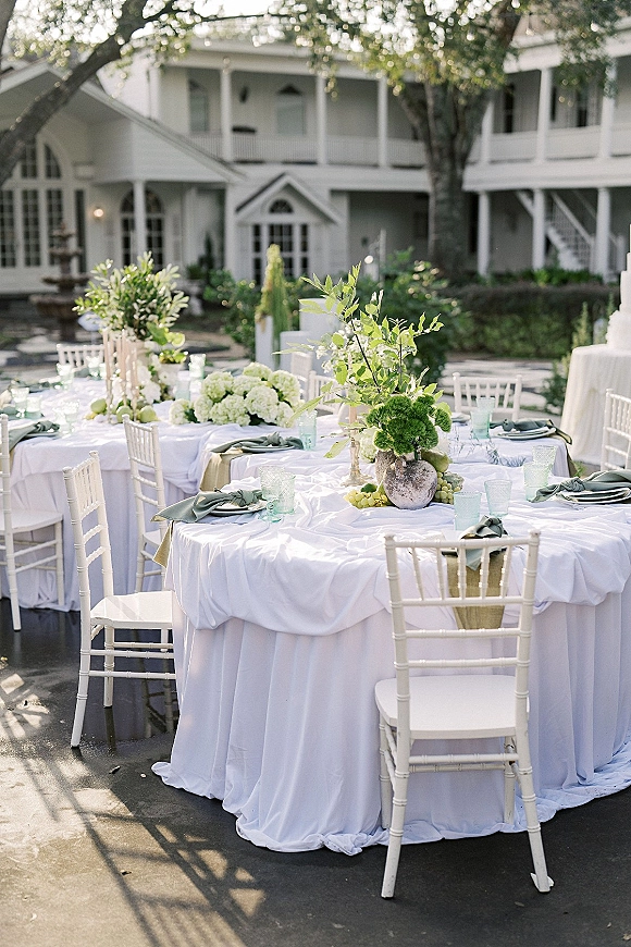 Reception tablescape with outdoor wedding reception details: round tables in white linens, greenery runners, floral centerpieces, taper candles on a patio