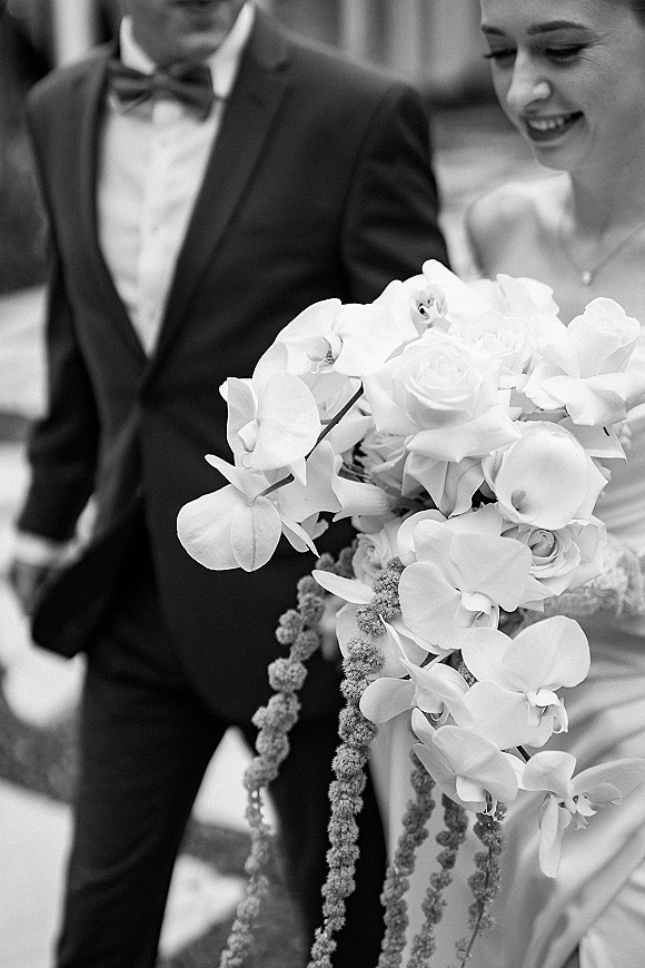 Bridal bouquet of white orchids and roses in a cascading design, held by a bride in a strapless dress beside groom on stone walkway