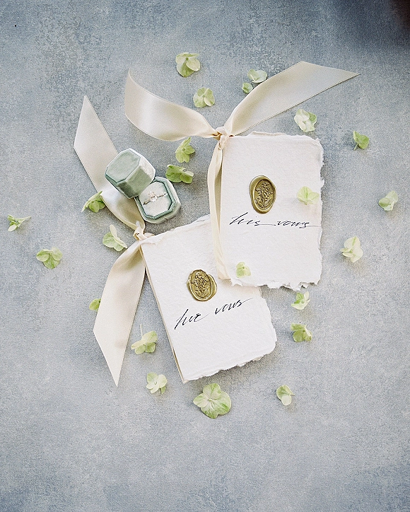 Vow books with silk ribbon ties and wax seals beside a ring box and engagement ring on a gray textured surface, scattered petals