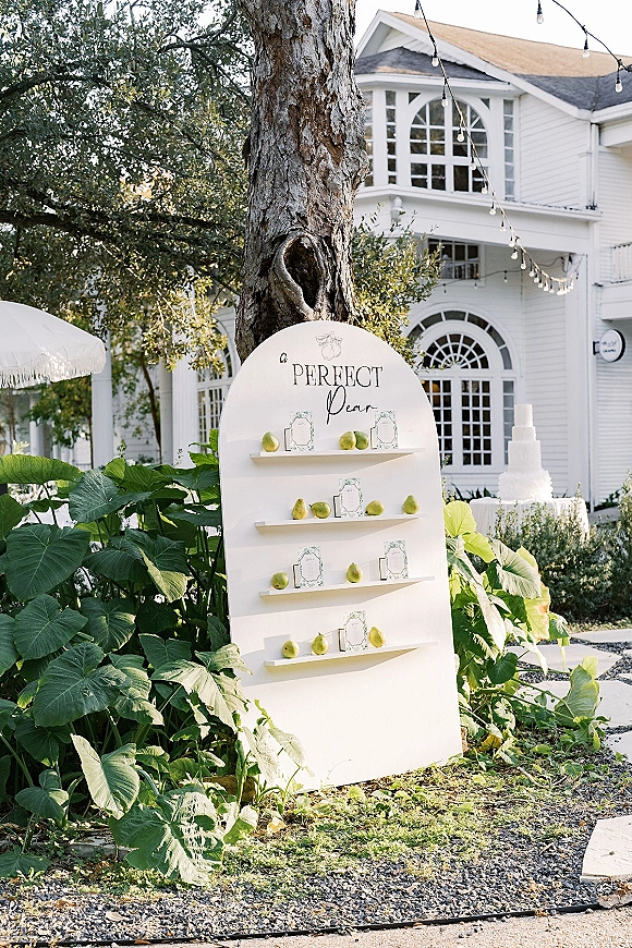 Seating chart display with arched seating chart board and calligraphy place cards on ledges, accented with pears and string lights outdoors