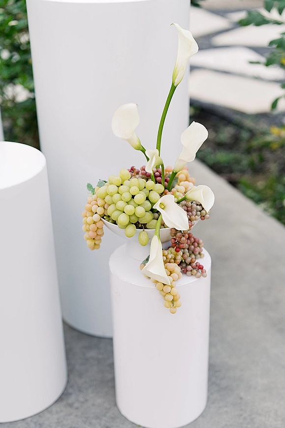 Wedding centerpiece with calla lily centerpiece and trailing grapes on a white pedestal stand, set along an outdoor garden walkway