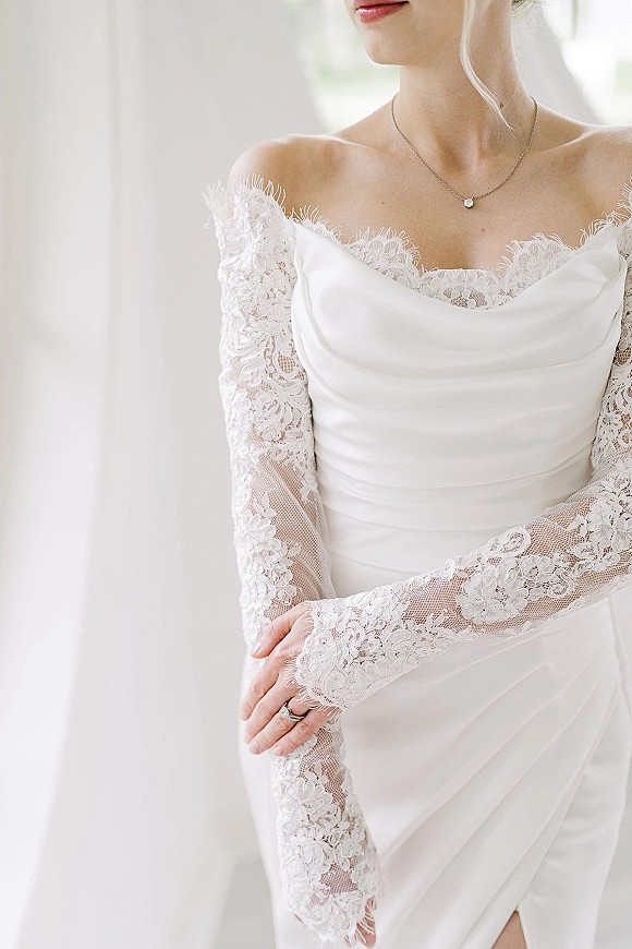 Wedding dress with off the shoulder neckline and lace sleeves, draped cowl bodice, pendant necklace and ring in window light