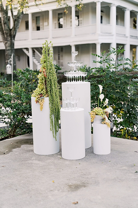 Champagne tower setup with wedding champagne tower of coupe glasses on white pedestal plinths, grapes and hanging amaranthus on a garden patio