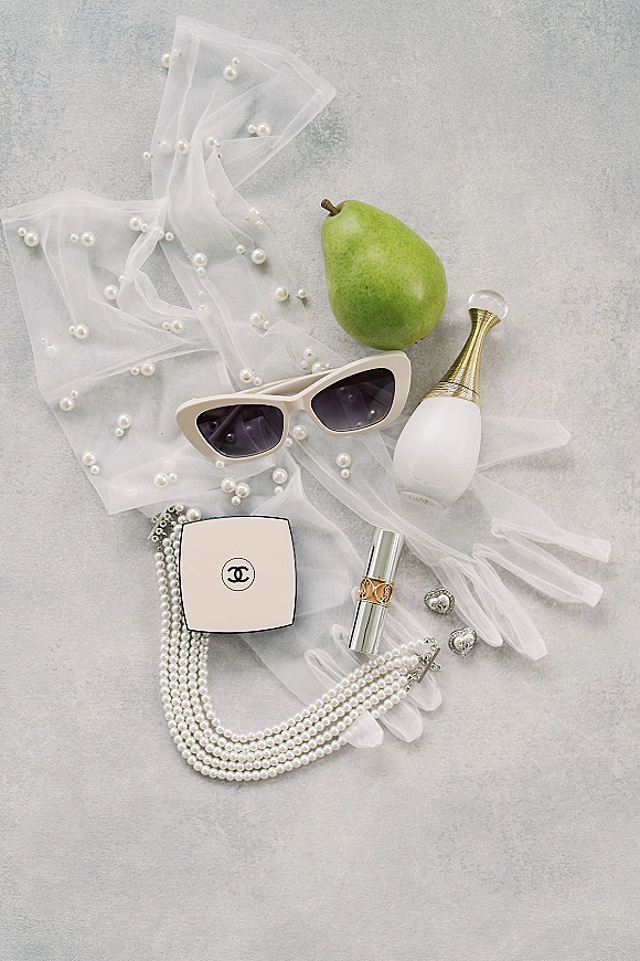 Bridal accessories flatlay with pearl necklace, veil, perfume bottle, lipstick, earrings, sunglasses, and green pear on gray stone surface