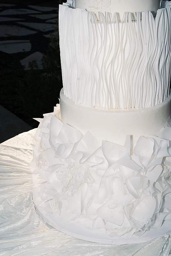 Wedding cake with modern wedding cake styling, all-white fondant tiers featuring textured panels and ruffled details on a white stand outdoors