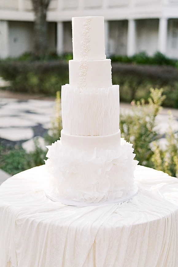 Wedding cake with textured white tiered design and sugar flower appliqué on a table with white cloth in an outdoor garden setting