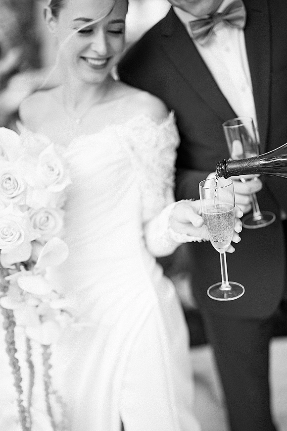 Wedding toast as bride and groom raise champagne flutes, bouquet and tuxedo bow tie visible against a softly blurred indoor backdrop