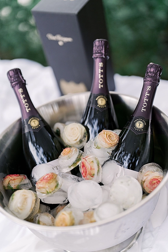 Champagne ice bucket with champagne bottles on ice, frozen rose ice cubes, and gift box beside a green upholstered sofa on white fabric