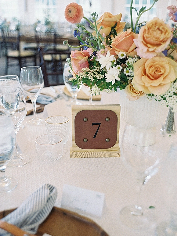 Reception tablescape with wedding floral centerpiece in a white ceramic vase, table number 7, place cards, and textured glass votives in window light