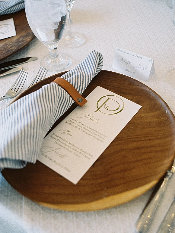 Wedding place setting with a wood charger plate, striped napkin, menu and place card, silver flatware, and glassware on white linens