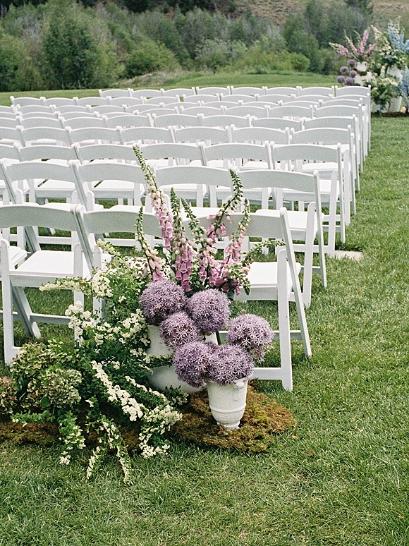 Ceremony seating with outdoor ceremony chairs lining an aisle, framed by urn vases with purple allium and mossy florals on a green lawn
