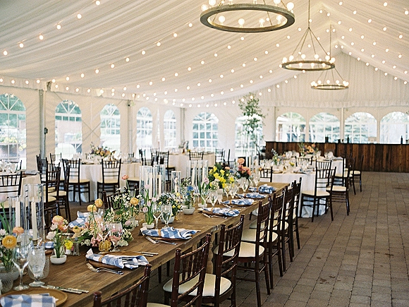 Reception tablescape in a wedding tent reception with blue napkins, wildflower centerpieces, taper candles, and string lights under draped ceiling