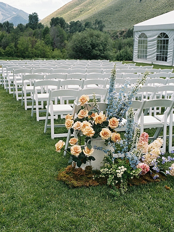 Ceremony seating with white folding chair rows and a moss-based floral arrangement at the aisle end on a lawn with mountains beyond