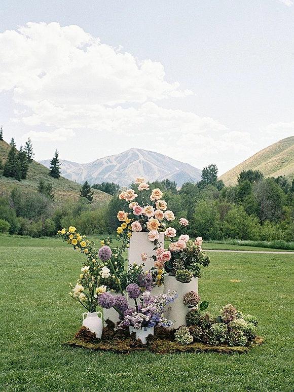 Ceremony floral arrangement of wedding altar flowers on white plinths with pastel roses, set against a mountain landscape and clouds