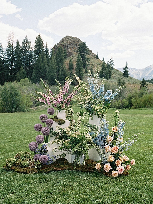 Wedding cake display with a tiered white wedding cake surrounded by pink roses and delphinium on white pedestals, set on a lawn with mountains beyond
