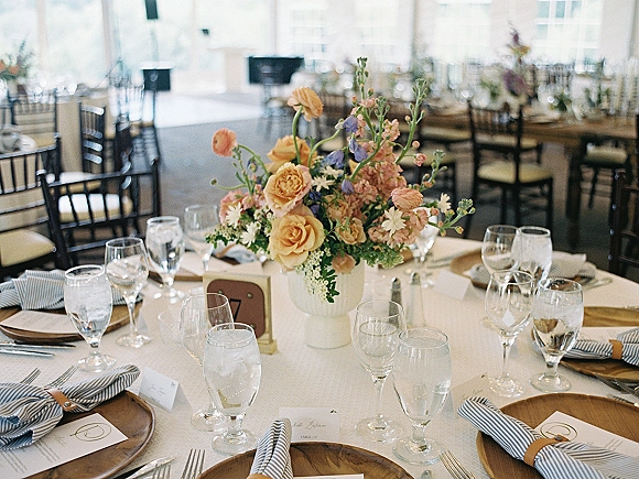 Reception tablescape with a wedding table centerpiece in a white vase, wood chargers, striped napkins, and place cards in an airy room with windows