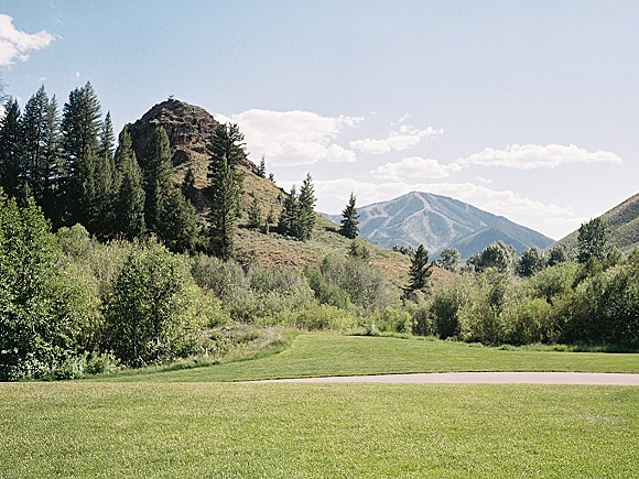 Mountain landscape with alpine valley scenery, pine trees bordering a grassy meadow beneath rugged peaks and blue sky with clouds
