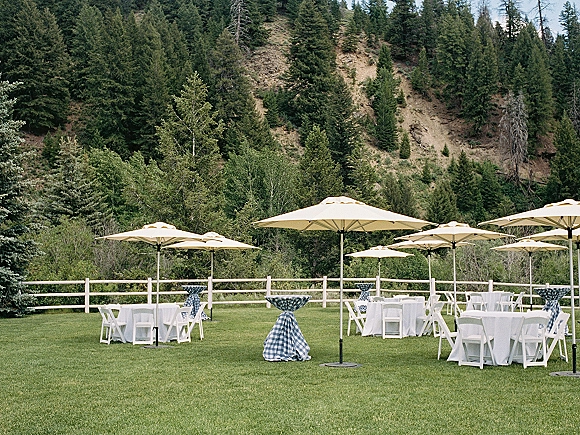 Outdoor reception setup with round tables, white tablecloths, and patio umbrellas on a grassy lawn beside a rustic wooden fence and trees