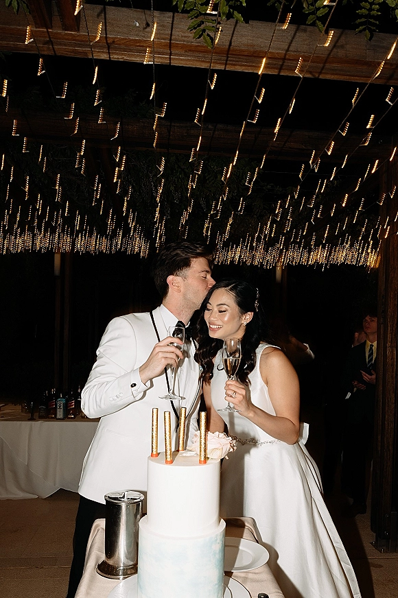 Wedding cake cutting as bride in a minimalist gown and groom in white jacket slice beside champagne flutes under pergola string lights at night