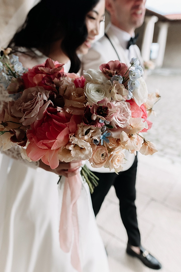 Bridal bouquet, colorful wedding bouquet of mixed coral and blush flowers with a blush ribbon, held by bride and groom on stone patio walkway
