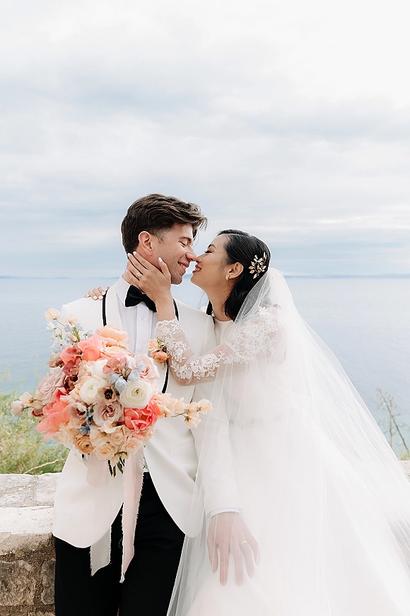 Wedding kiss portrait of bride kissing groom as she holds his face, bouquet and veil visible beside a stone wall with ocean beyond