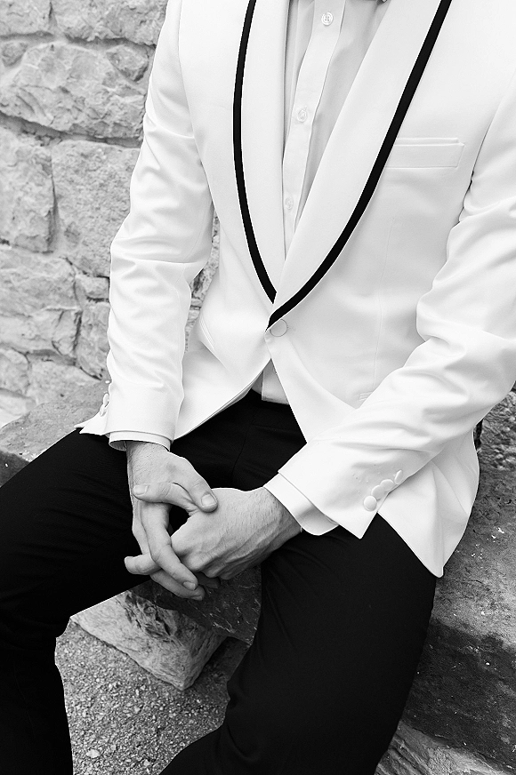 Groom portrait in a white tuxedo jacket with black shawl lapel trim, seated with hands clasped by a stone wall and bench