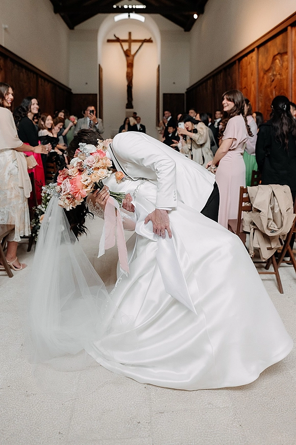 Wedding kiss portrait of groom dipping bride in a church aisle, veil and satin lace gown flowing as guests cheer by wooden pews