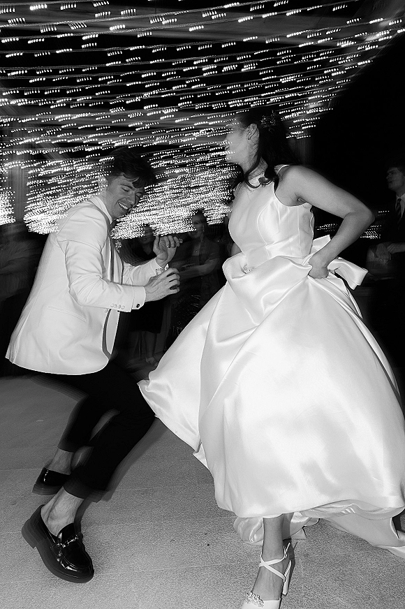 Wedding dance moment as bride twirls in a bow-back dress beside groom in white tuxedo jacket under string lights on a crowded dance floor