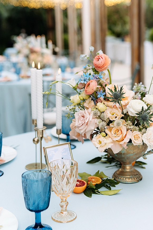 Reception tablescape with wedding floral centerpiece in a pedestal bowl, brass taper candles, blue goblets, citrus slices, and string lights on a patio