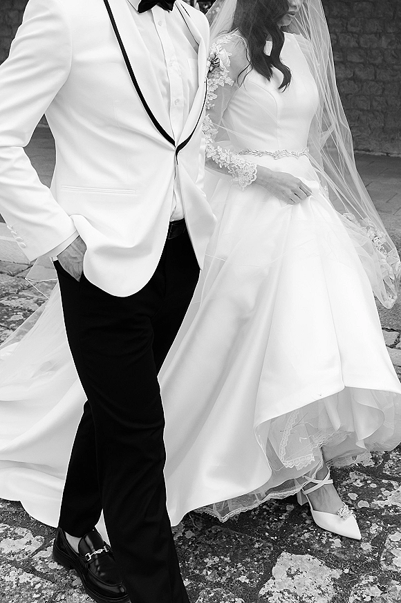 Couple portrait in a black and white wedding portrait style, bride in lace sleeves and cathedral veil walking with groom in white dinner jacket by a stone wall