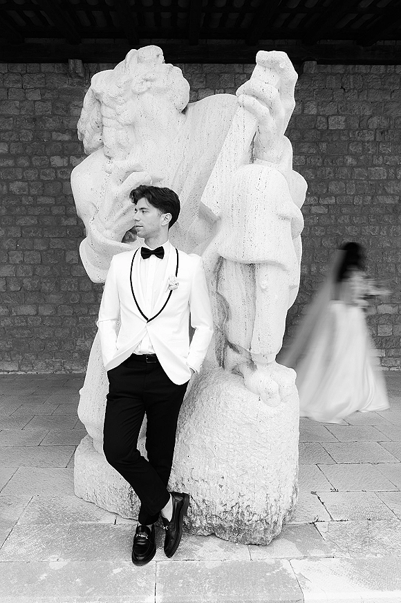 Groom portrait in a white tuxedo jacket with boutonniere, hands in pockets by a stone statue and wall as bride blurs behind him
