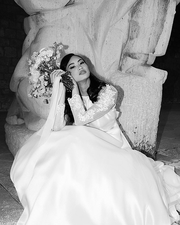 Bridal portrait of a bride in a lace-sleeve wedding dress sitting on a stone floor, holding a bouquet with ribbon by a stone sculpture