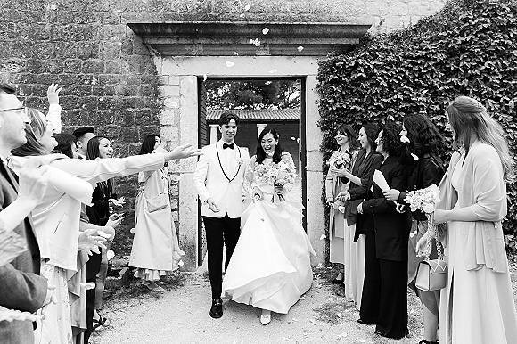 Wedding recessional as newlyweds walking out through a stone doorway, bride in full skirt gown with bouquet, guests tossing petals in courtyard
